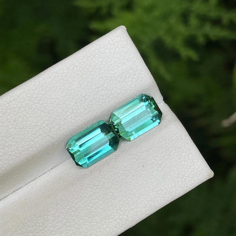 May include: Two rectangular, teal-colored gemstones with a bright, reflective surface. The gems are set on a white surface, with a blurred green background. The gems have a rectangular cut with visible facets.