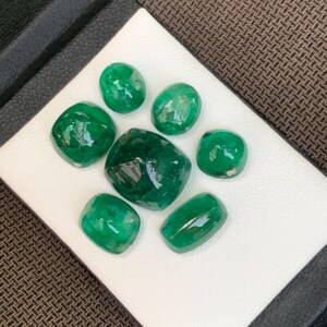 May include: A collection of seven loose, polished emerald gemstones of varying sizes and shapes, including oval, cushion, and rounded cuts. Each stone displays a deep, vibrant green color with natural internal inclusions and a smooth, reflective surface.