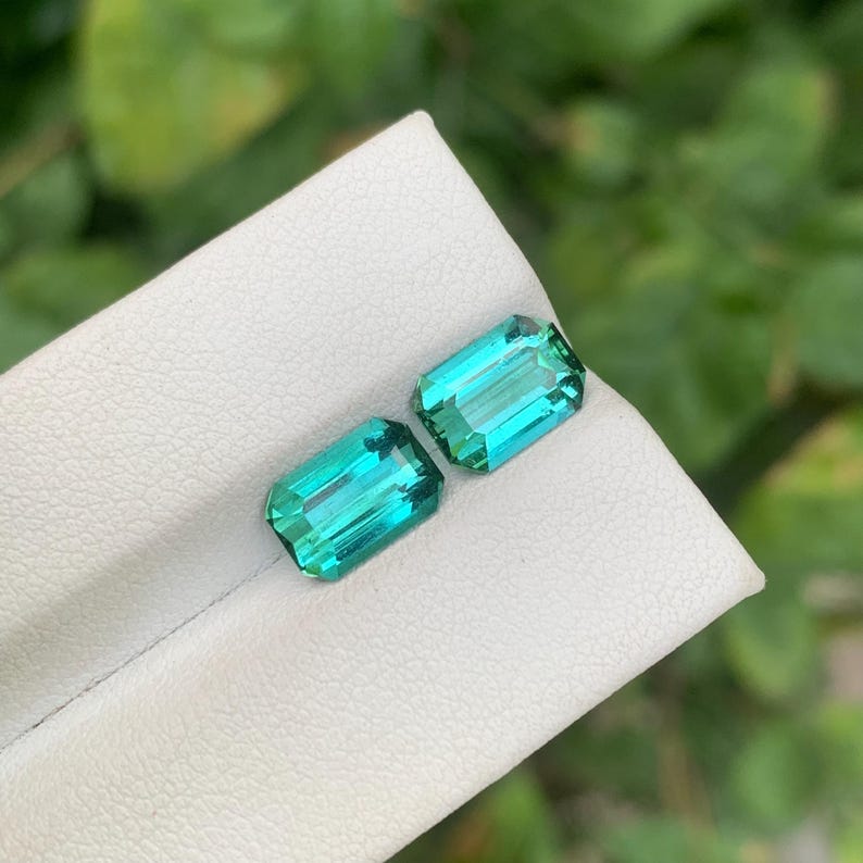 May include: Two rectangular, emerald-cut gemstones with a vibrant teal color. The gems are displayed on a white surface, with a blurred green background. The facets of the stones reflect light, showcasing their clarity and brilliance. These gems are likely for jewelry.