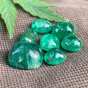 May include: A collection of seven polished, cabochon-cut emerald green gemstones with smooth, rounded surfaces and varying sizes, displaying natural internal inclusions and light reflections, arranged together in a cluster.