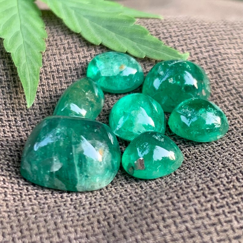 May include: A collection of seven polished, cabochon-cut emerald green gemstones with smooth, rounded surfaces and varying sizes, displaying natural internal inclusions and light reflections, arranged together in a cluster.