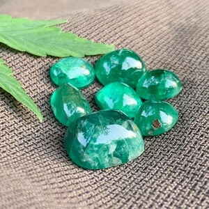 May include: A collection of seven loose, polished emerald gemstones with a smooth, rounded cabochon cut. The stones exhibit a vibrant, translucent green color with visible internal inclusions and reflective surfaces, arranged in a cluster.