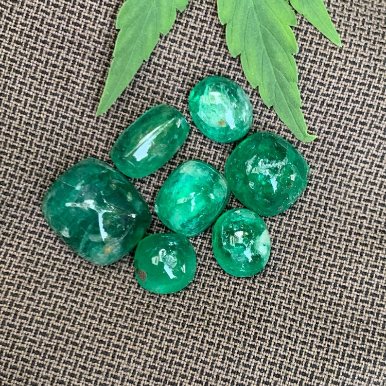 May include: A collection of seven loose, polished emerald gemstones in various shapes and sizes, displaying a vibrant, translucent green color with natural internal inclusions, arranged together near a green leaf on a textured brown surface.