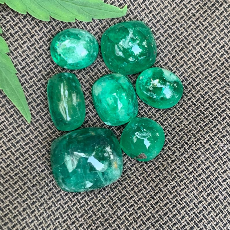 May include: A collection of seven polished, translucent green emerald gemstones with smooth, rounded surfaces and varying shapes, including oval and cushion cuts, displaying natural internal inclusions and vibrant emerald green color tones.