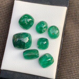 May include: Seven loose, polished emerald gemstones of varying sizes and shapes, including oval, cushion, and rectangular cuts. Each stone displays a deep, vibrant green color with natural internal inclusions and a smooth, reflective surface.