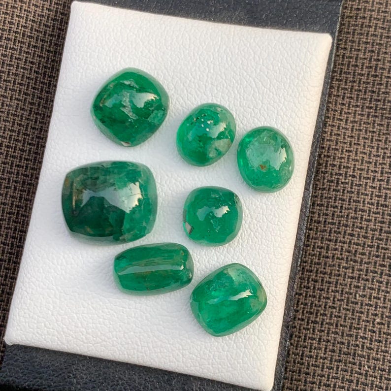 May include: Seven loose, polished emerald gemstones of varying sizes and shapes, including oval, cushion, and rectangular cuts. Each stone displays a deep, vibrant green color with natural internal inclusions and a smooth, reflective surface.