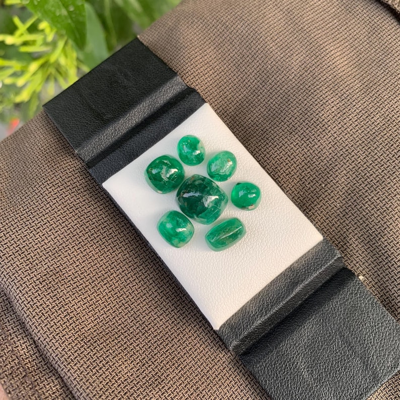 May include: A collection of seven loose, polished green gemstones of varying sizes and shapes arranged on a white rectangular display card, which is secured by two black leather-like bands against a textured brown fabric background.