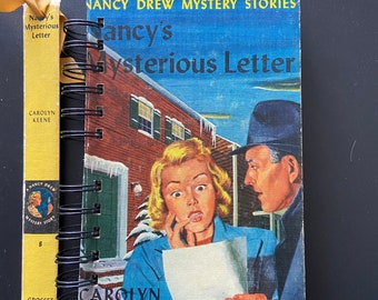 Puede incluir: Una portada de libro vintage de Nancy Drew, un misterio, con una mujer leyendo una carta con un hombre mirando por encima de su hombro. El título del libro es "Nancy's Mysterious Letter" de Carolyn Keene.