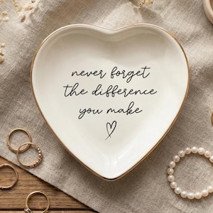 May include: A heart-shaped white ceramic trinket dish with a gold-painted rim. The center features black cursive text that reads, never forget the difference you make, accompanied by a small hand-drawn heart symbol below the lettering.
