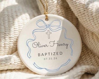 May include: A white ceramic baptism ornament with a light blue ribbon and cross design. The ornament has the name "Oliver Henry" and the word "BAPTIZED" with a date. A beige string is attached for hanging.