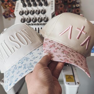 Può includere: Due cappellini da baseball con disegni ricamati. Uno è bianco con "WILSON" a lettere grandi e una visiera floreale blu. L'altro è beige con lettere ricamate rosa e una visiera floreale rosa. I cappellini sono tenuti in mano.