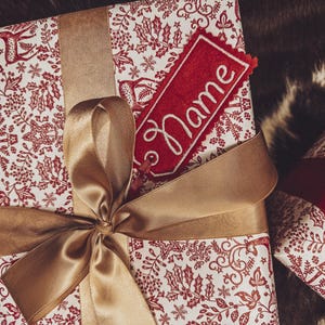 May include: A wrapped gift box with a festive red and white floral pattern, tied with a gold satin ribbon and bow. A red tag with the word "Name" in white script is attached. The gift is on a textured surface.