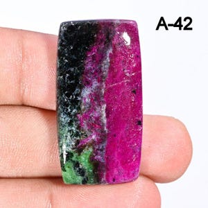 Peut inclure: Une pierre précieuse rectangulaire à surface polie. La pierre présente un dégradé de couleurs saisissant, avec des sections noires, rose foncé, vertes et blanches. Le texte "A-42" est visible dans le coin supérieur droit.