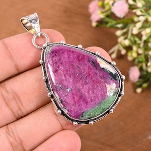 May include: A teardrop-shaped pendant featuring a large, polished ruby zoisite gemstone with vibrant magenta and green inclusions. The stone is set in a silver bezel with small decorative metal beads along the edge and a textured silver bail.