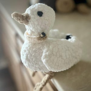 May include: A handmade white crochet duck with a beige beak, feet, and collar. The duck has black eyes and is sitting on a surface. This soft sculpture is a charming decorative item.