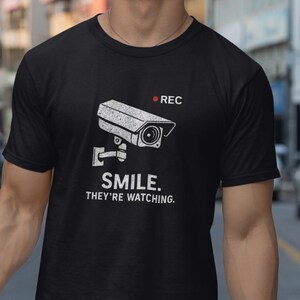 May include: Black t-shirt with a white graphic of a security camera and the text "SMILE. THEY'RE WATCHING." The shirt has a crew neck and short sleeves. The person is wearing white shorts and is smiling.