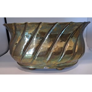 May include: An oval brass planter featuring a polished metallic finish with a distinct diagonal ribbed or fluted pattern along the exterior walls. The vessel has a scalloped top edge and is supported by small, rounded feet at the base.