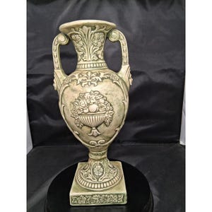 May include: A pale green ceramic vase featuring two handles and a square base. The body is decorated with a relief of a fruit bowl surrounded by floral patterns and scrollwork. The surface has a weathered finish with darker green accents.