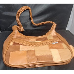 May include: A hobo-style handbag constructed from a patchwork of various tan and brown leather squares. The bag features a braided leather shoulder strap and a matching braided leather trim detail across the front exterior panel with a zipper.