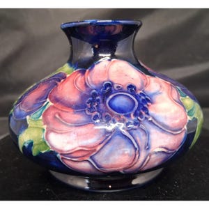 May include: A squat, bulbous ceramic vase featuring a deep cobalt blue base colour. The surface is decorated with large, raised pink flower blossoms with dark blue centres and subtle green foliage accents, all finished with a glossy, crackled glaze.