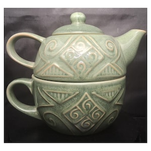 May include: A sage green ceramic tea set for one, featuring a stackable teapot with a lid and a matching teacup. Both pieces are decorated with embossed geometric patterns, including central diamonds with swirls and triangular accents.