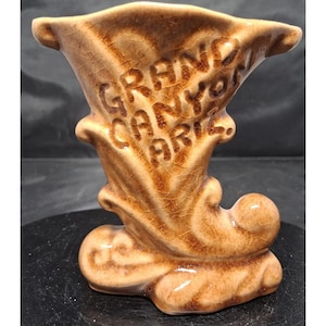 May include: A brown ceramic vase with a glossy crackle glaze finish. The front features embossed text that reads GRAND CANYON ARIZ. The vase has a flared top opening and a curved, sculptural base with decorative scroll-like details on the sides.