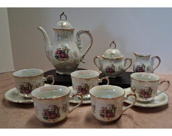 Puede incluir: Un juego de té de porcelana con acabado iridiscente y bordes dorados. El conjunto incluye una tetera con tapa, un azucarero con tapa, una jarra de leche y cuatro tazas con platillos, cada una decorada con una escena colorida de figuras.