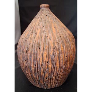 May include: A teardrop-shaped ceramic vase with a textured, bark-like surface in shades of brown and orange. The exterior features vertical grooves and small, scattered circular perforations throughout the body, leading to a narrow neck.