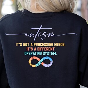 May include: Black sweatshirt with the word "autism" in cursive script. Below the word, the text reads "IT'S NOT A PROCESSING ERROR. IT'S A DIFFERENT OPERATING SYSTEM." An infinity symbol made of puzzle pieces is below the text.