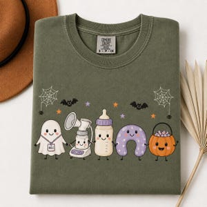 May include: An olive green crewneck t-shirt featuring a graphic of a ghost with an IBCLC badge, a breast pump, a baby bottle, a purple nursing pillow, and a jack-o'-lantern bucket, all with faces, surrounded by bats, spiderwebs, and stars.
