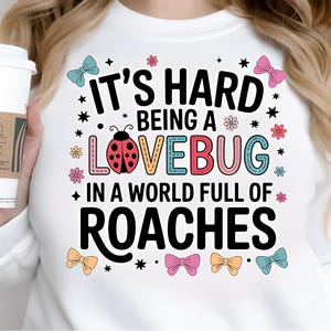 May include: White sweatshirt with the text "It's Hard Being a Lovebug in a World Full of Roaches." The design includes a ladybug, bows, and stars. A person is holding a white coffee cup with a brown sleeve.