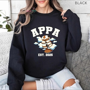 May include: Black sweatshirt with the word "APPA" in white block letters above a cartoon character with brown and white fur, and the text "EST. 2025" below. The character is surrounded by white clouds.