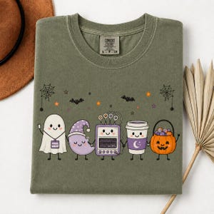 May include: An olive green crewneck t-shirt featuring a graphic of a ghost, a crescent moon, a medical heart monitor, a coffee cup, and a jack-o'-lantern holding hands. The ghost wears a badge labeled RPSGT. Small bats, stars, and spiderwebs surround them.