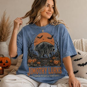 May include: A blue short-sleeve t-shirt featuring a graphic of a raccoon in a graveyard with pumpkins, bats, and a large orange moon. The text reads JIMOTHY LURKS IN PUMPKIN PATCHES & GRAVEYARDS in orange lettering across the bottom of the design.