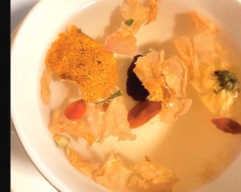 May include: A white bowl filled with clear liquid and various dried ingredients. The ingredients include orange peel, dried flowers, and small red berries. The liquid has a light amber hue, and the bowl has a gold rim.