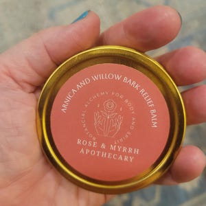 May include: A round, gold-colored tin with a pink label. The label reads "ARNICA AND WILLOW BARK RELIEF BALM" and "ROSE & MYRRH APOTHECARY". The text is in a serif font. The tin is held in a hand.