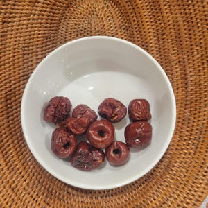 May include: A white ceramic bowl containing dried red dates. The dates are a deep reddish-brown colour with a wrinkled texture. The bowl sits on a woven, light brown surface.