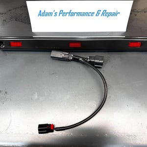 May include: A black automotive light bar with three red rectangular lights. A black wire harness with connectors extends from the light bar. The text "Adam's Performance & Repair" is visible in the background.