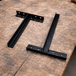 May include: Two black metal shelf brackets in a T-shape, each with pre-drilled holes for mounting. The brackets are displayed on a weathered wooden surface, showcasing their industrial design and potential for home decor.