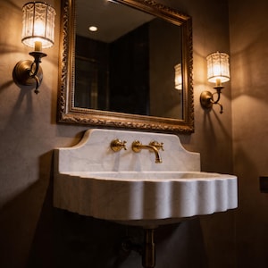 May include: A wall-mounted white marble sink with a scalloped front edge and gold-toned faucet fixtures. Above the sink hangs a rectangular mirror with an ornate gold frame, flanked by two cylindrical glass wall sconces emitting warm light.