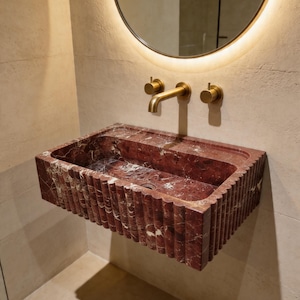 May include: A red marble rectangular sink with a fluted design, mounted on a beige wall. The sink has a gold-finished faucet and two knobs. A round mirror with warm lighting is above the sink.