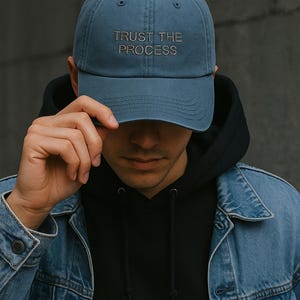 Peut inclure: Casquette de baseball bleue avec l'inscription "TRUST THE PROCESS" brodée en or. La casquette est portée avec un sweat à capuche noir et une veste en jean.
