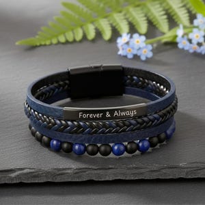 May include: A multi-layered bracelet with a black magnetic clasp. The bracelet features a blue leather band, a braided black band, and a band with black and blue beads. A black metal plate is engraved with the words "Forever & Always".