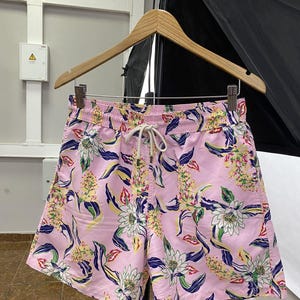 May include: Pink floral print shorts with a drawstring waist. The shorts feature a vibrant pattern of white, yellow, and blue flowers and leaves. The shorts are hanging on a wooden hanger.