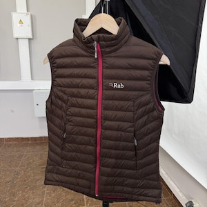 May include: A brown puffer vest with a high collar and a red zipper. The vest has the brand name "Rab" in white on the chest. It is hanging on a wooden hanger.