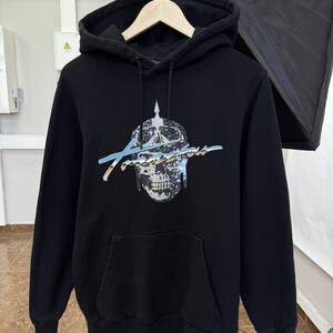 May include: A black hooded sweatshirt featuring a graphic print of a skull wearing a spiked helmet with a galaxy-like pattern. The word Trapstar is written in a stylized, metallic-effect script across the center, with a small yellow tooth detail.