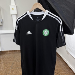 May include: A black short-sleeved jersey with a white V-neck collar and white stripes on the shoulders. The shirt features a white Adidas logo on the right chest and the green and white Celtic Football Club crest on the left chest.