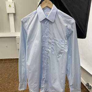 May include: A light blue long-sleeve button-down shirt with a subtle vertical pinstripe pattern. The shirt features a classic collar, a single chest pocket on the left side, and white buttons running down the center placket and on the cuffs.