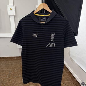 May include: A black and dark gray horizontal striped polo shirt featuring a white New Balance logo on the right chest and a white Liverpool Football Club crest with the text L.F.C. and AXA printed below it on the left chest.