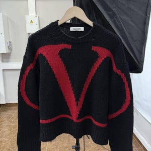 May include: Black knitted jumper featuring a bold red "V" motif. The crew neck sweater has long sleeves and the brand name "VALENTINO" is visible on the label. The garment is displayed on a wooden hanger.